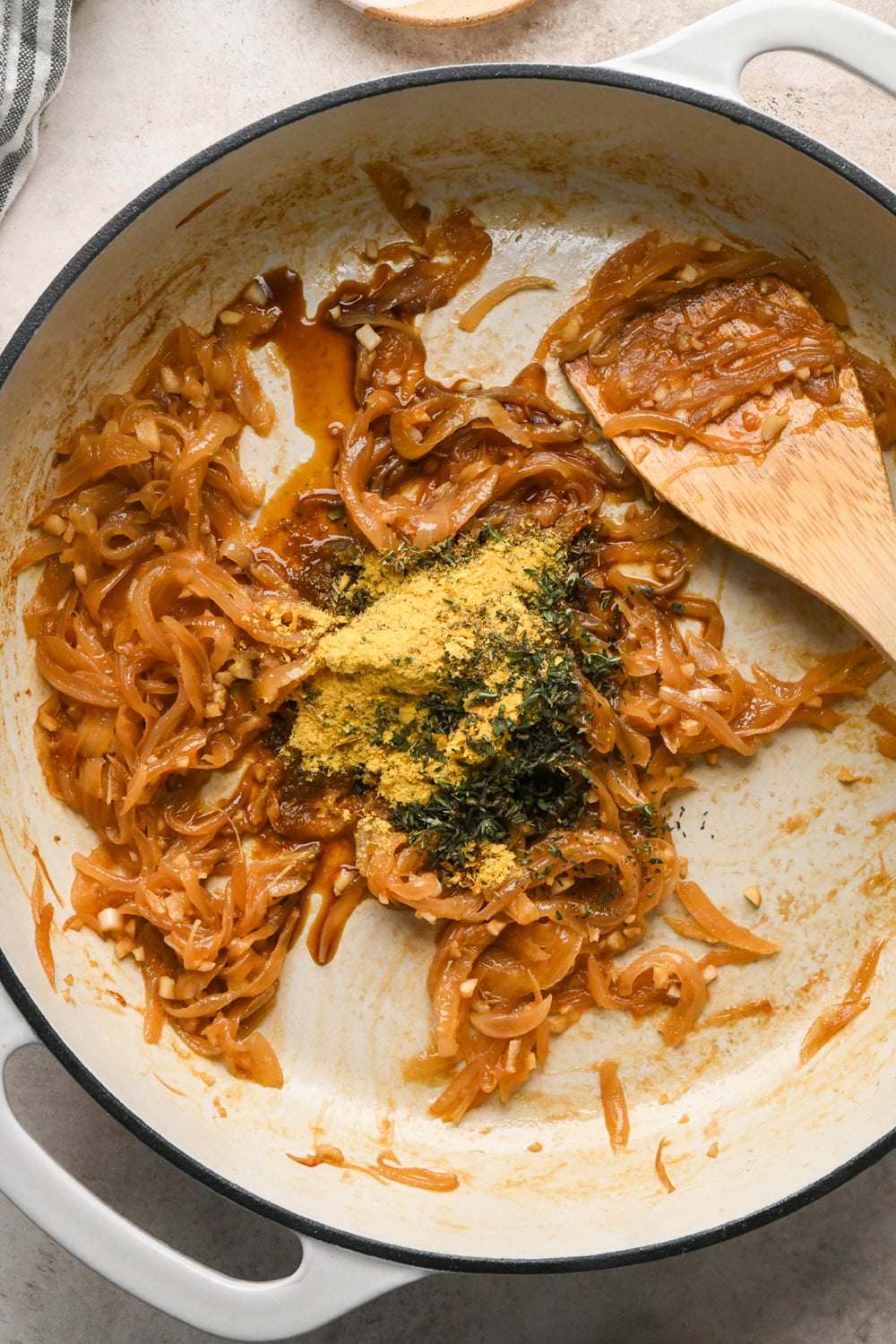 Caramelized onions in a white pan, topped with a mound of nutritional yeast, chopped thyme, and coconut aminos, with a wooden spatula resting on the side.