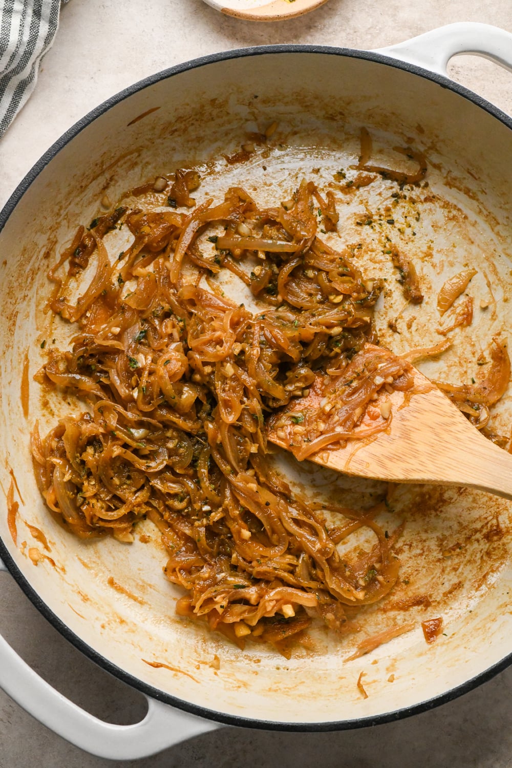 Caramelized onions in a white pan, after nutritional yeast, chopped thyme, and coconut aminos have been stirred into the mixture, with a wooden spatula resting on the side.