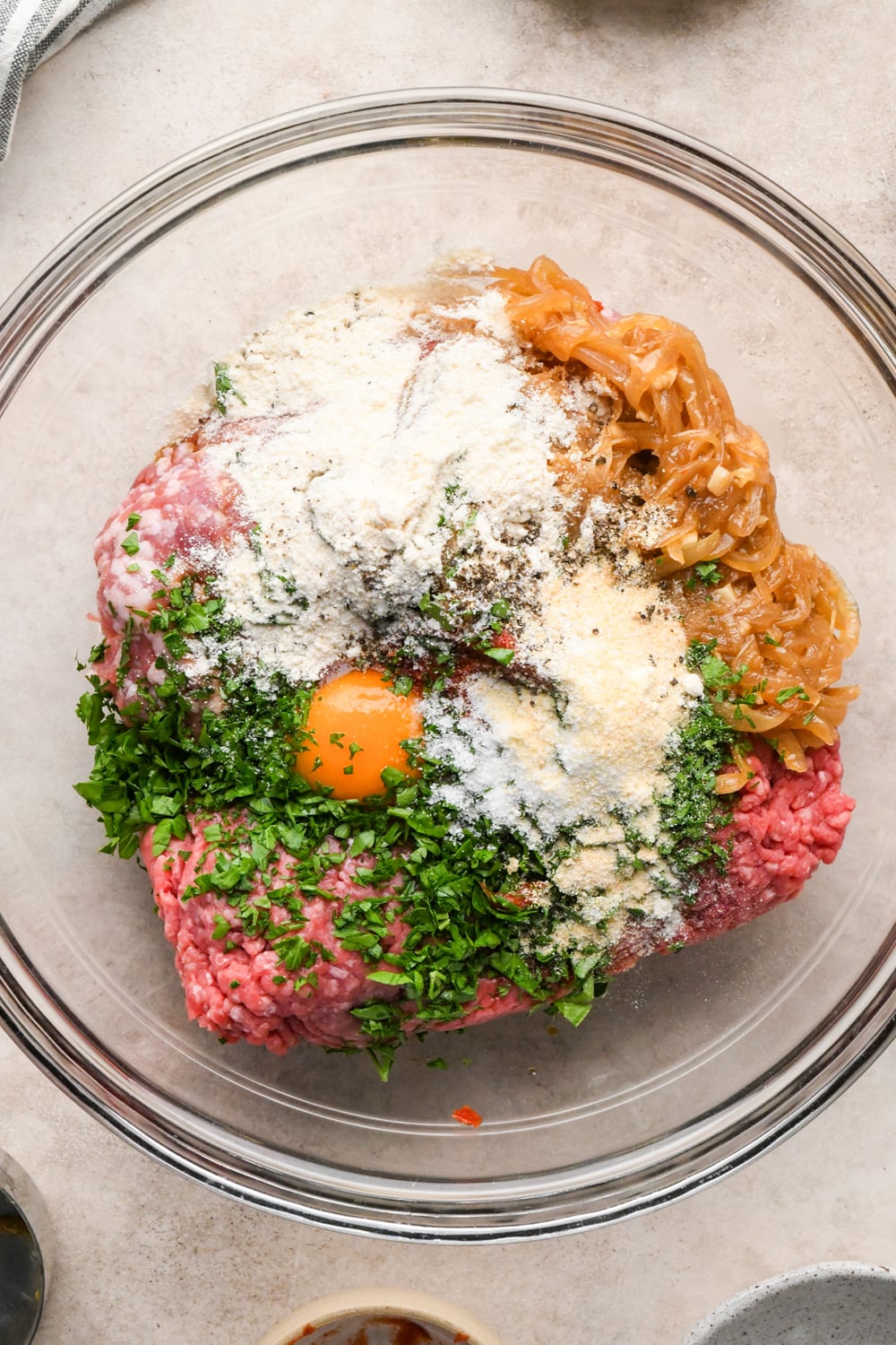 A glass bowl containing raw ground meat, a cracked egg, coconut flour, dried spices, sautéed onions, and fresh chopped herbs.