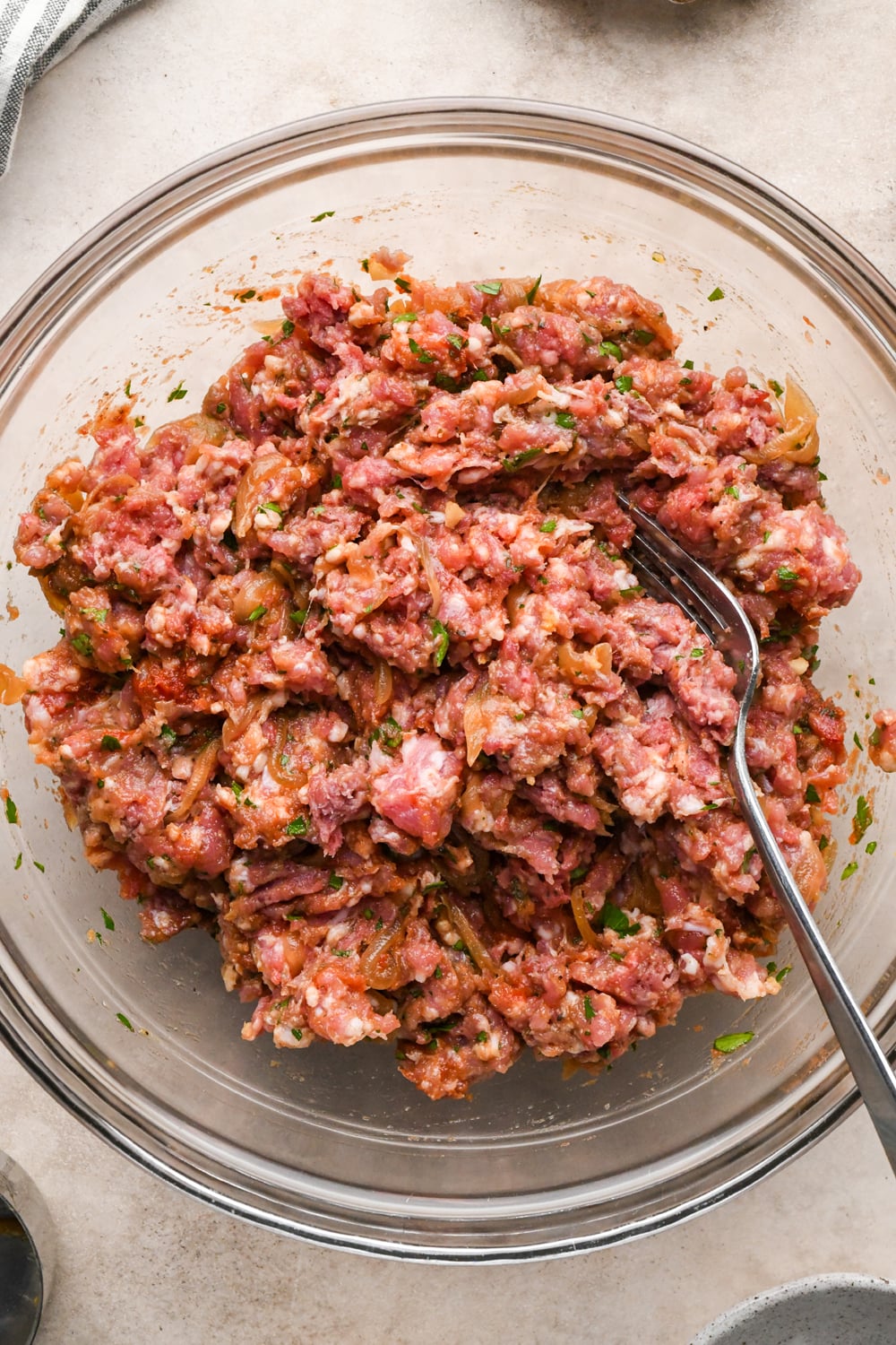 A glass bowl filled with raw ground meat mixed with herbs and sautéed onions, with a fork resting inside.