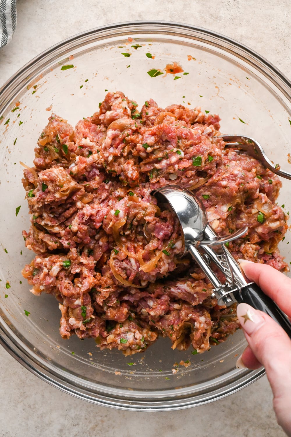 A glass bowl filled with raw ground meat mixed with herbs and sautéed onions, with a ice cream scoop dipping into the mixture.