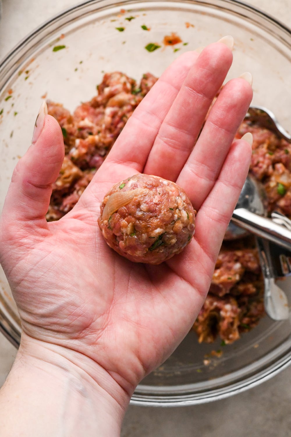 A hand holding a raw meatball above a glass bowl filled with seasoned ground meat.