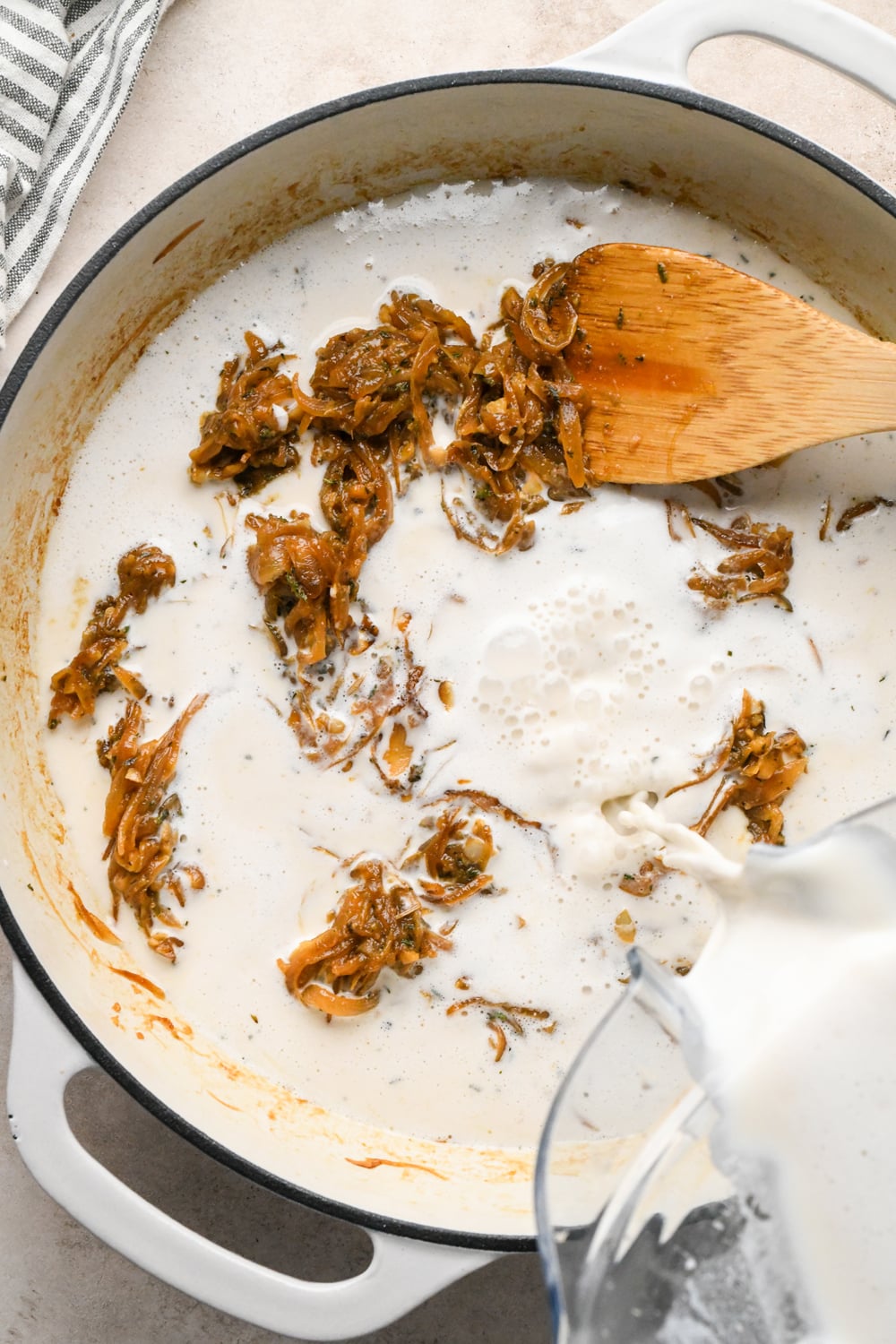 Cashew cream sauce being poured into a pan of caramelized onions and herbs, with a wooden spoon resting inside.