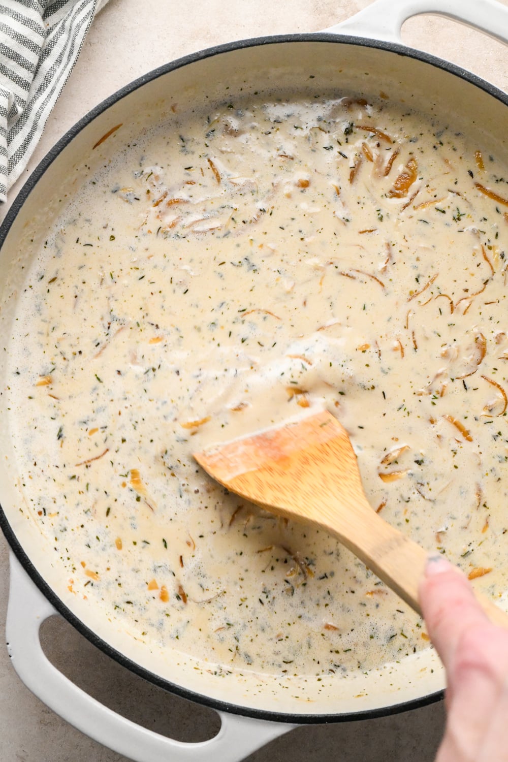 A wooden spoon stirs a creamy, herb-flecked sauce with sautéed onions in a large white skillet.