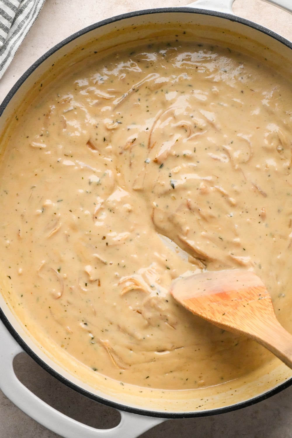 A wooden spoon stirs creamy gravy with herbs in a white enameled cast iron pan.