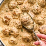 A silver spoon lifts a caramelized onion meatball coated in creamy gravy from a skillet filled with meatballs in the same sauce.