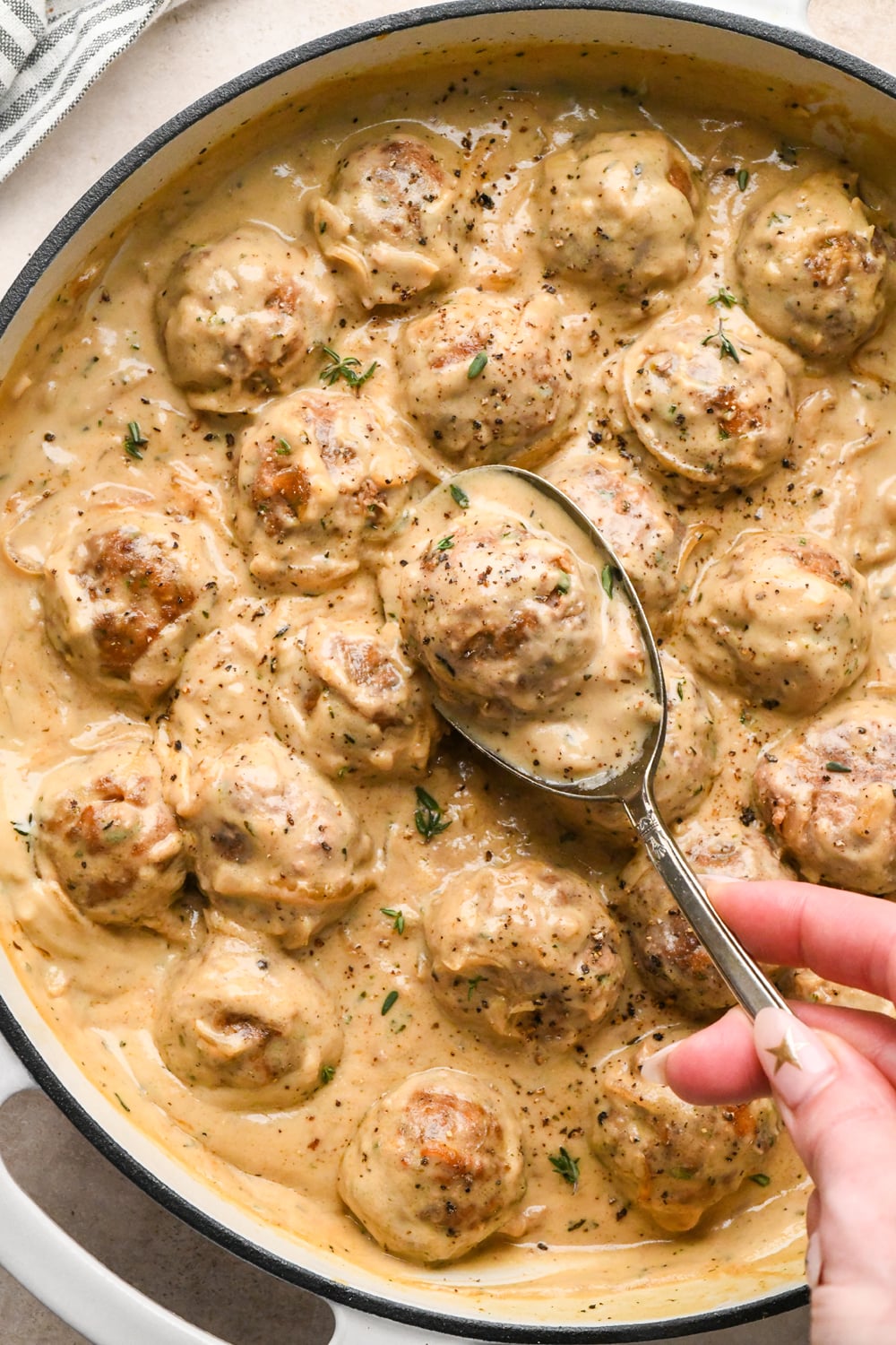 A silver spoon lifts a caramelized onion meatball coated in creamy gravy from a skillet filled with meatballs in the same sauce.
