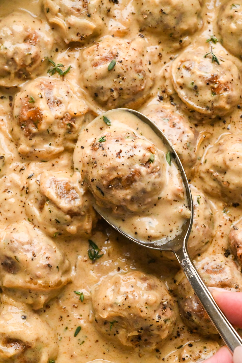 A silver spoon lifts a caramelized onion meatball coated in creamy gravy from a skillet filled with meatballs in the same sauce.