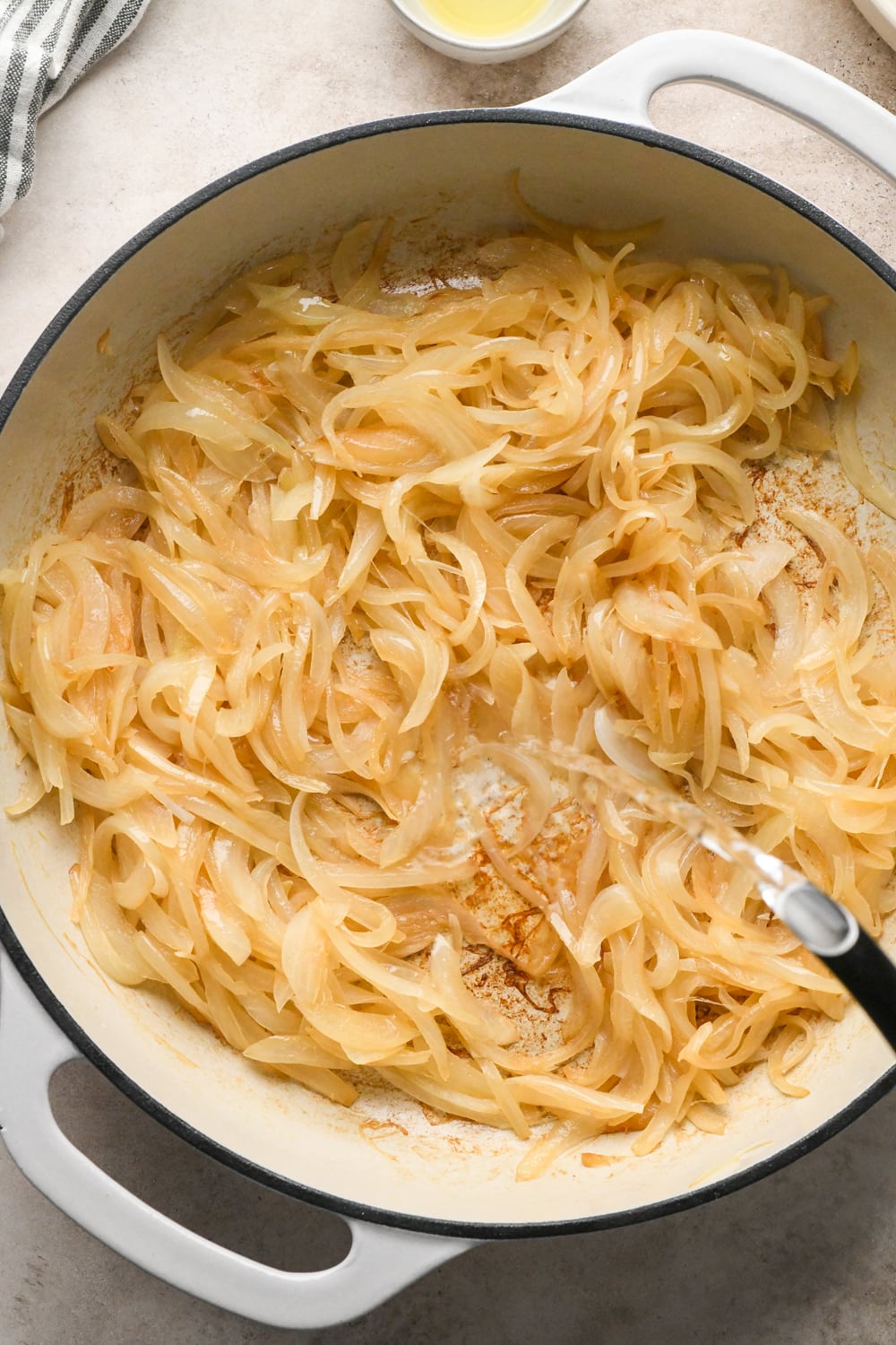 Sliced onions that are slightly caramelized, with a black tea kettle pouring some water into the skillet, with a wooden spatula.