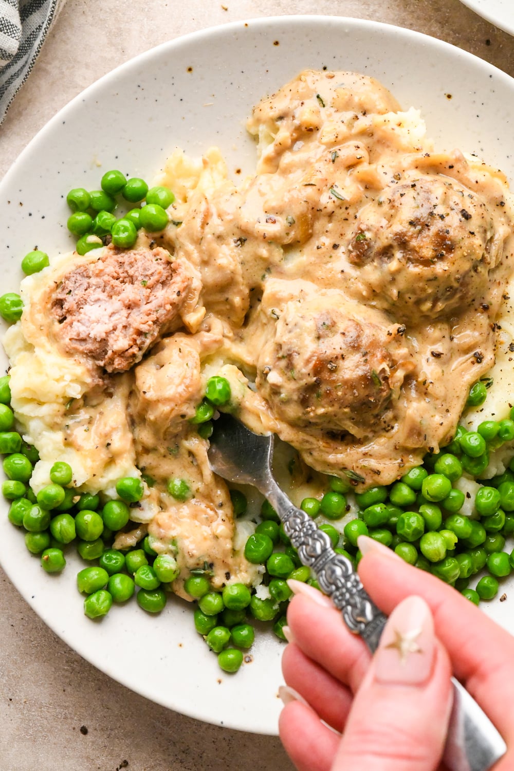 A fork cuts into a caramelized onion meatball covered in rich and creamy gravy, served with mashed potatoes and green peas on a white speckled plate.