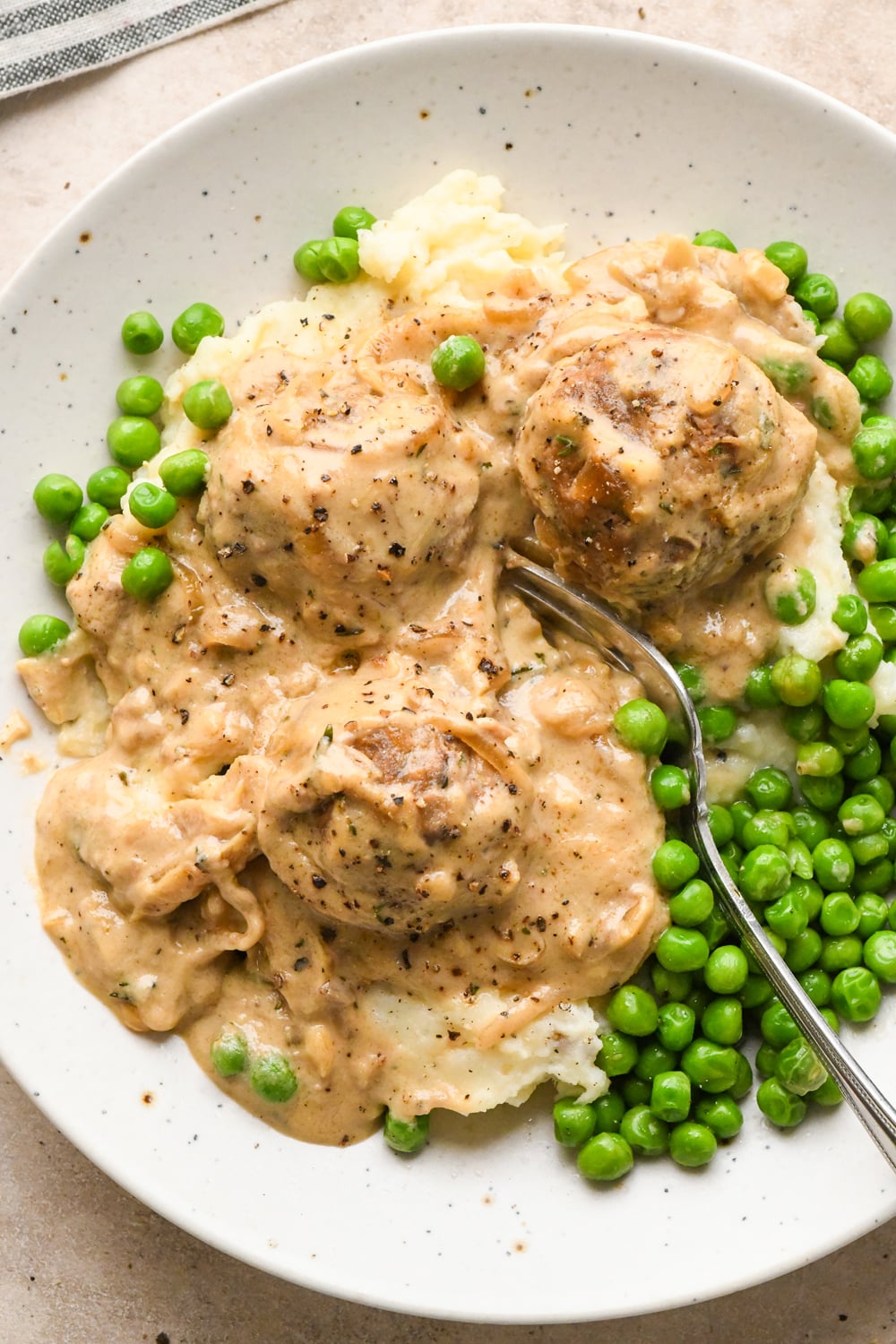Dairy-free caramelized onion meatballs in gravy served over mashed potatoes with a side of green peas, with a fork resting on the plate.
