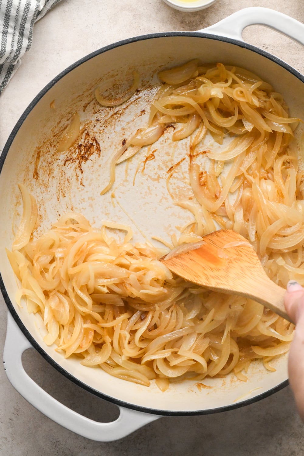 Sliced onions that are slightly caramelized, cooking in a white enameled cast iron skillet with a wooden spatula stirring them around to release the stuck on bits using water that has just been poured into the skillet.
