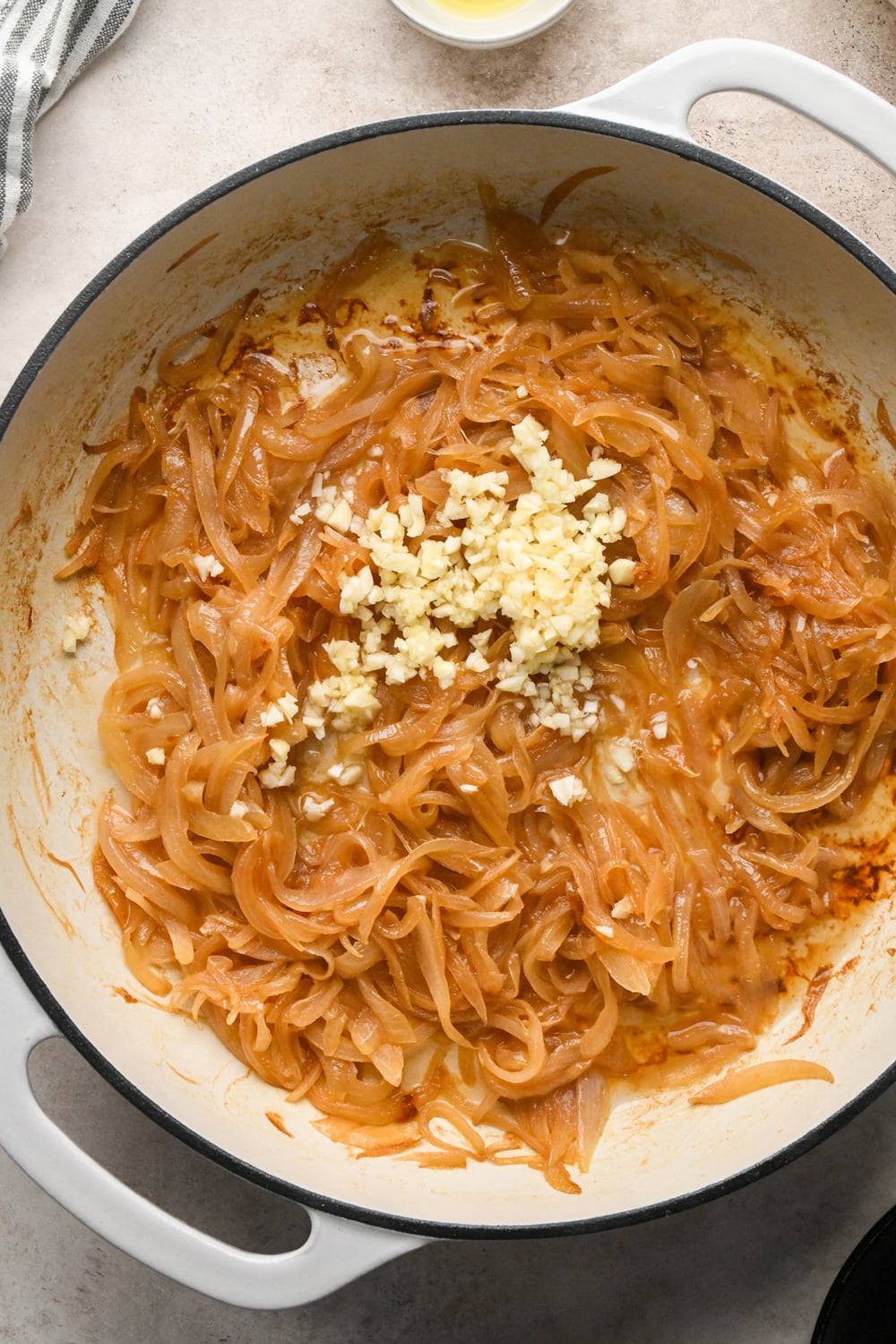 Deeply caramelized sliced white onions in a white cast iron skillet, with raw garlic added.